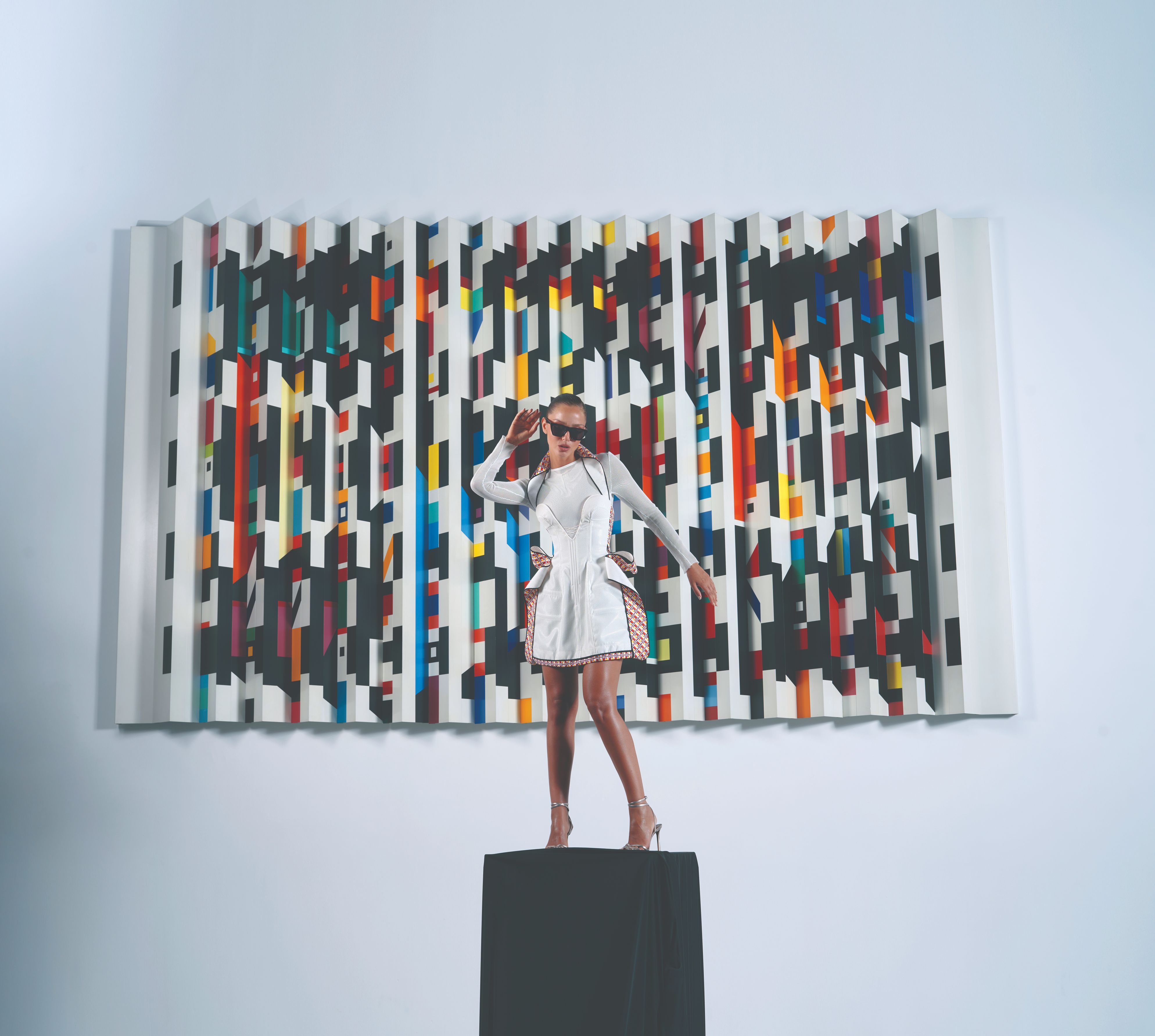 דוגמנית מהפקת אופנה העומדת על במה שחורה ומאחוריה ציור צבעוני 